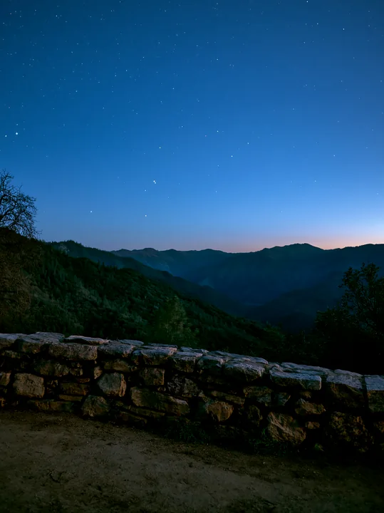 Stars fill the night sky above a mountain valley, with a stone wall in the foreground.
