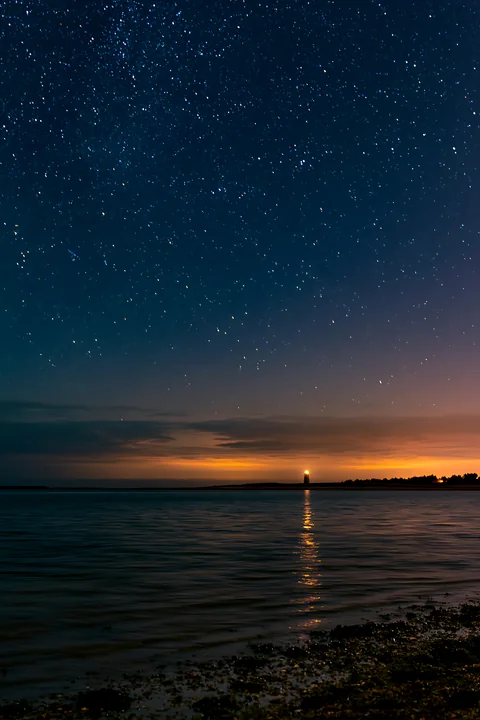 Stars fill the night sky over a calm ocean, with a lighthouse visible on the horizon reflecting light on the water.