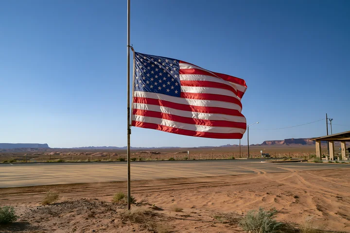 The American flag flying in strong wind stands prominently beside a road in a desert landscape.