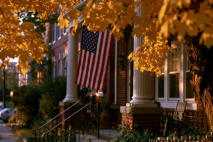 The American flag hanging from the porch adds character to the home, surrounded by vibrant autumn leaves.