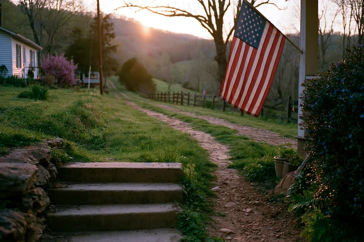The American flag on rural road hangs from a porch as the sun sets over the hills.
