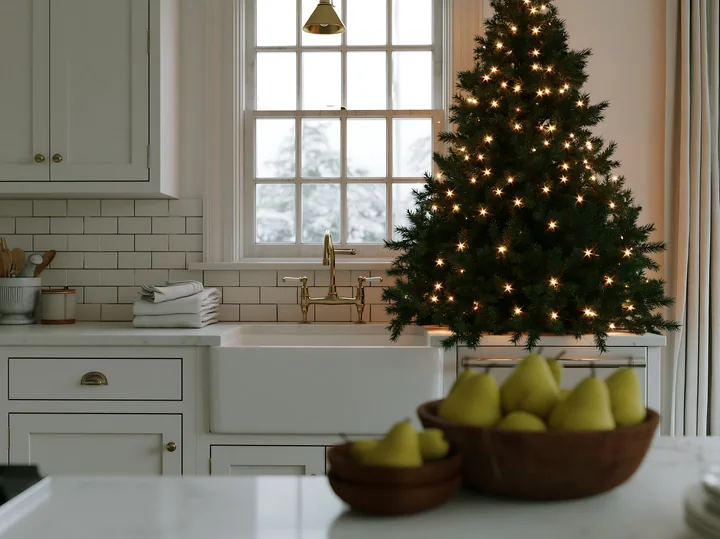 The scene shows a luxury kitchen decorated for the holiday season, featuring an illuminated Christmas tree near a window, natural light shining in.