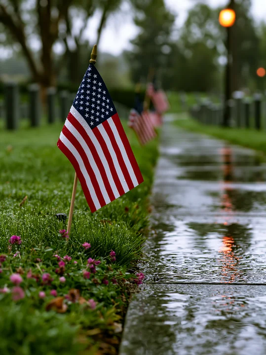 The scene shows small American flags placed on the grass along a wet pathway in a veterans cemetery.