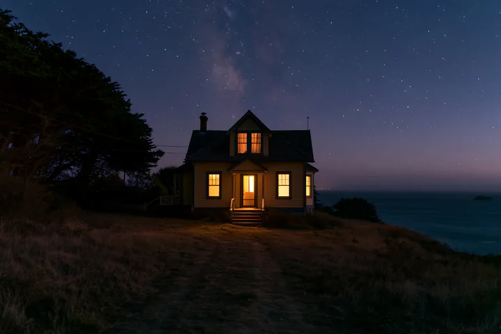 The villa sits on a hillside overlooking the ocean, with warm light coming from the windows, contrasting against the dark night filled with stars.