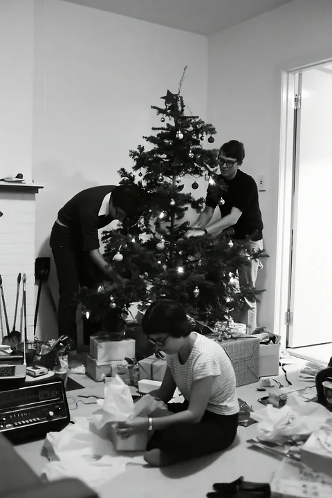 Three adults arrange wrapped gifts beneath a decorated Christmas tree in a home.