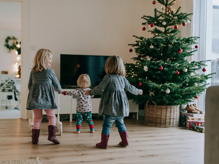 Three children dance joyfully around a decorated Christmas tree in a cozy living room.