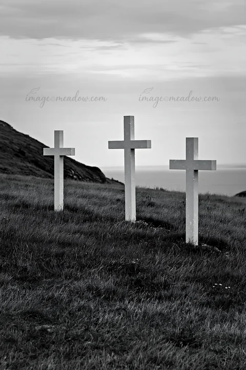 Three Christian crosses stand in a quiet meadow. The crosses are placed on grassy land, overlooking the sea. The scene has a simple and natural look, under a cl