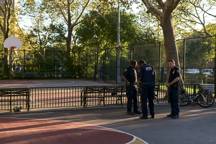Three police officers are examining an area in a basketball court located in a public park during daylight hours.