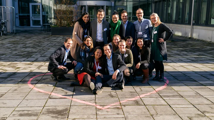 Twelve smiling colleagues pose inside a pink heart-shaped chalk outline on a paved courtyard outside an office building.