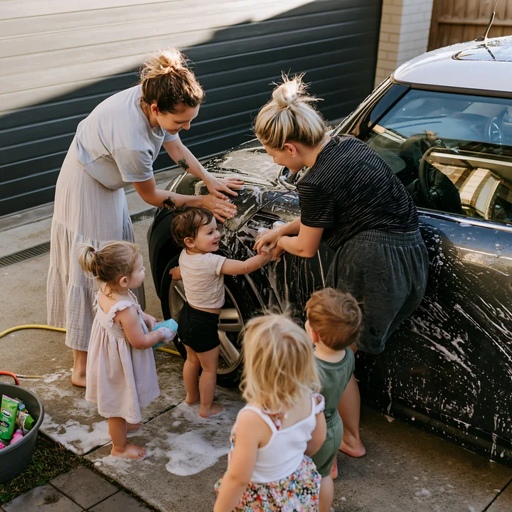 Two adults and four children wash a soapy car together in a residential driveway.