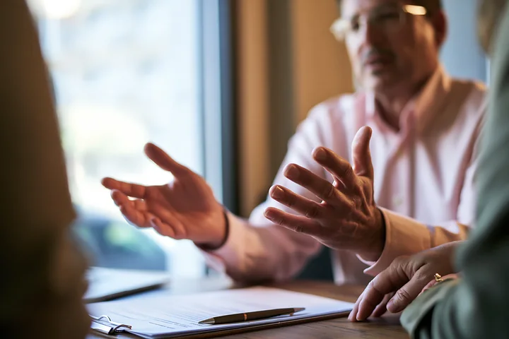 Two businessmen discuss paperwork across a desk in a warmly lit office.