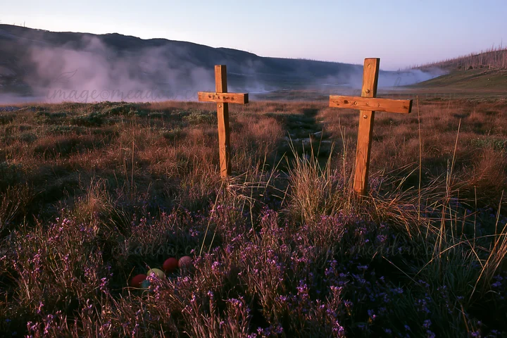 The scene shows two wooden Christian crosses standing in a grassy meadow near a thermal spring. The sun rises, casting light on the area surrounded by mist and