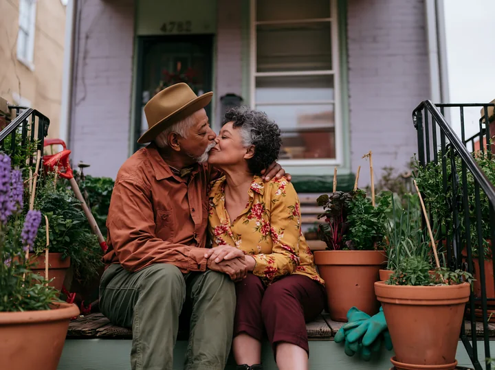 Two elderly partners share a kiss while sitting on their porch surrounded by potted plants.