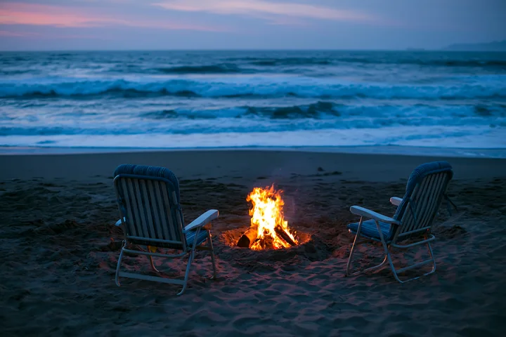 Two empty beach chairs face a glowing bonfire on the sandy shore with ocean waves at dusk.