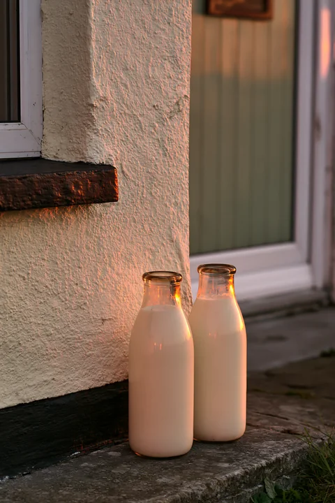 Two glass milk bottles rest on a stone doorstep as the sun rises, casting warm light against a textured wall.