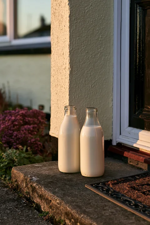Two glass milk bottles stand on a doorstep as the sun rises, casting soft light on the scene.