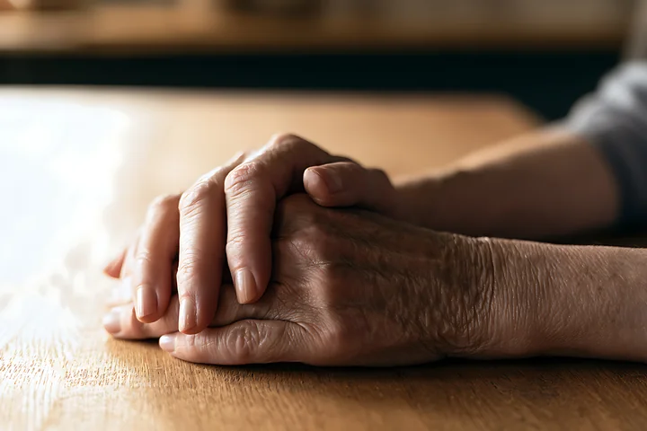 Two hands are intertwined and resting on a wooden table in warm, soft light.