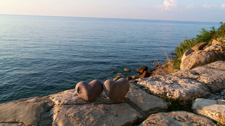 Two heart-shaped stones rest on sunlit coastal rocks beside calm blue ocean water.