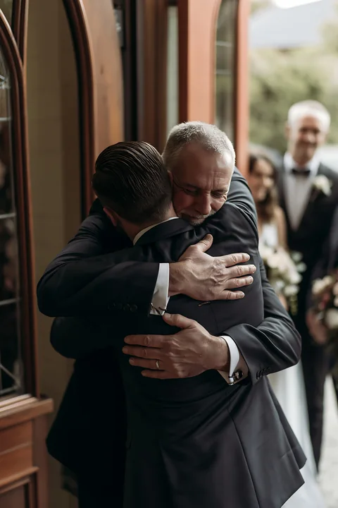 Two men in formal suits share an emotional embrace during a wedding ceremony near a doorway.