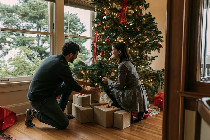 Two parents place wrapped gifts beneath a decorated Christmas tree in a warmly lit home.