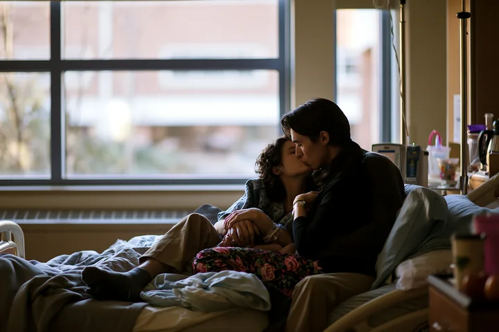 Two partners embrace and kiss beside a hospital bed in a softly lit room.