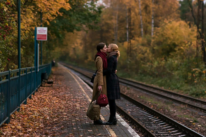 Two partners kiss beside railway tracks on a leaf-covered autumn train platform.