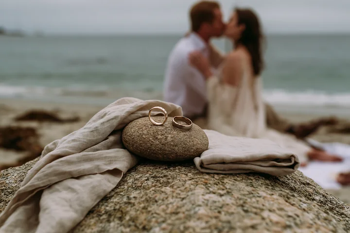 Two people kiss beside wedding rings resting on a rock at the beach.