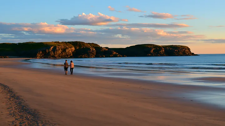 Two people walk along a sandy beach near a rocky peninsula as the sun sets in the background.