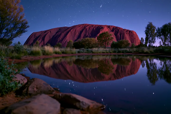 Uluru rises prominently against a star-filled sky as its reflection appears in a tranquil water body.