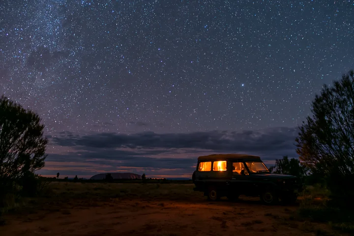 Uluru sits beneath a star-filled night sky, with an illuminated off-road vehicle in the desert foreground.