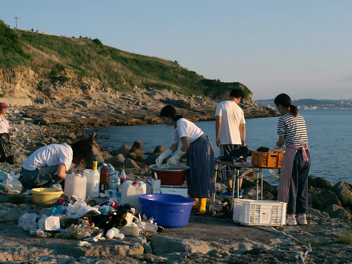 Volunteers clean up plastic waste along a rocky coastline during sunset.