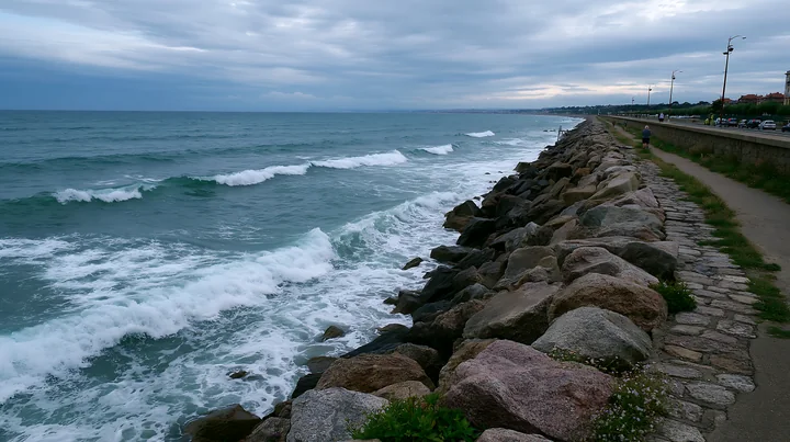 Waves break against a rocky sea wall beside a coastal road under a cloudy sky.