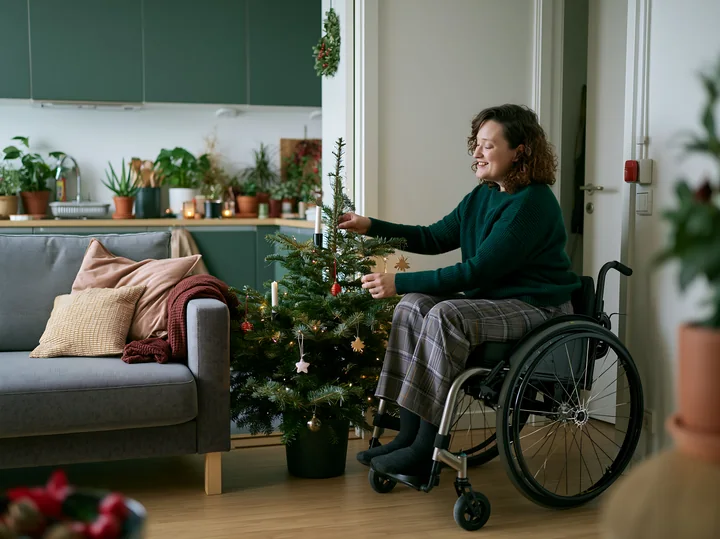 Woman in a wheelchair decorates a Christmas tree in a plant-filled apartment living room.