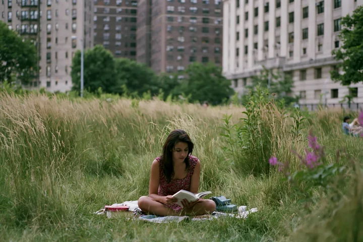 Woman sitting in tall grass and reading a book in an urban meadow surrounded by apartment buildings.