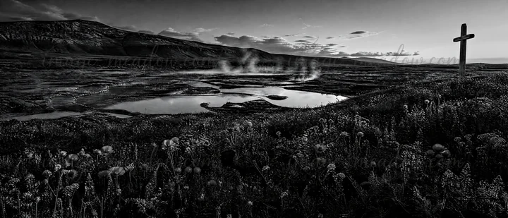 A wooden Christian cross stands in a meadow by hot springs. Steam rises from the ground, and the landscape features mountains and scattered vegetation. This sce